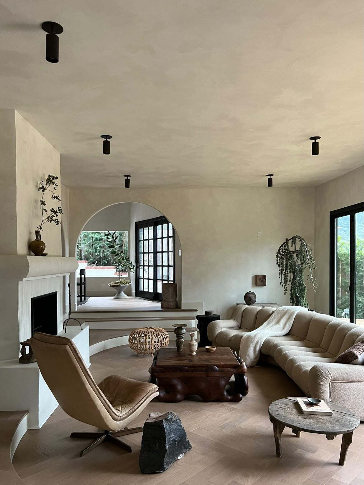A modern living room with neutral tones features a curved beige sofa, tan lounge chair, wooden coffee table, plants, and large windows. Cove Adjustable Spot by Long Made Co. and black framed doors enhance the space's airy elegance.