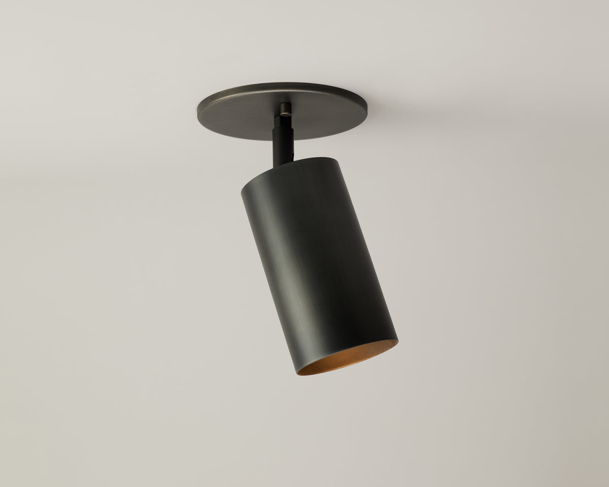The Cove Adjustable Spot by Long Made Co. is a modern black cylindrical ceiling light from the Cove Series, mounted on a round base and angled downward for focused, minimalist illumination against a white ceiling.