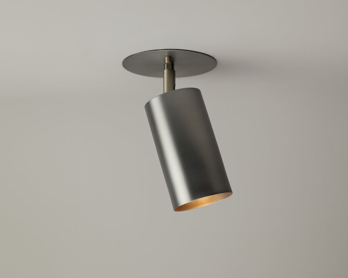 The Cove Adjustable Spot by Long Made Co. is a modern, cylindrical solid steel downlight with a brushed finish, mounted on a round plate and angled downward—ideal for those seeking minimalist spotlights.