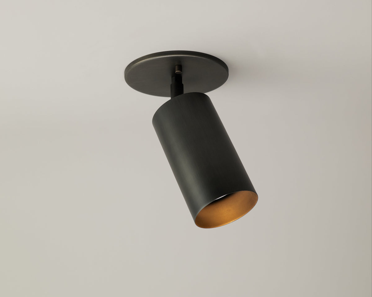 The Cove Adjustable Spot by Long Made Co. is a modern black cylindrical steel downlight from the Cove Series, mounted on a round base against a white ceiling, with its minimalist spotlight angled downward.