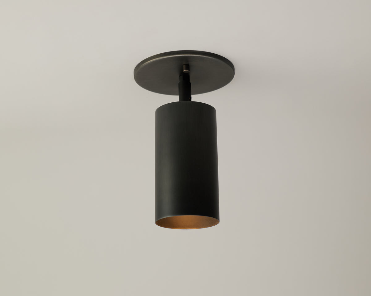 The Cove Adjustable Spot by Long Made Co. is a modern, cylindrical black ceiling light with a matte finish and gold-toned interior, mounted on a round base against a white ceiling.