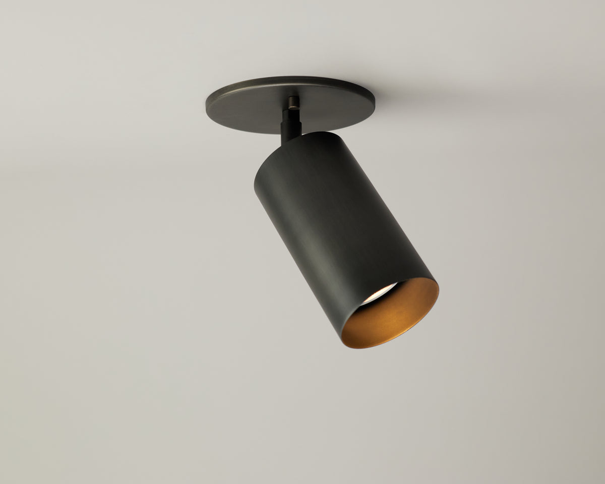 The Long Made Co. Cove Adjustable Spot, a black cylindrical solid steel downlight, is mounted on a plain white ceiling and angled downward to provide minimalist spotlight illumination.