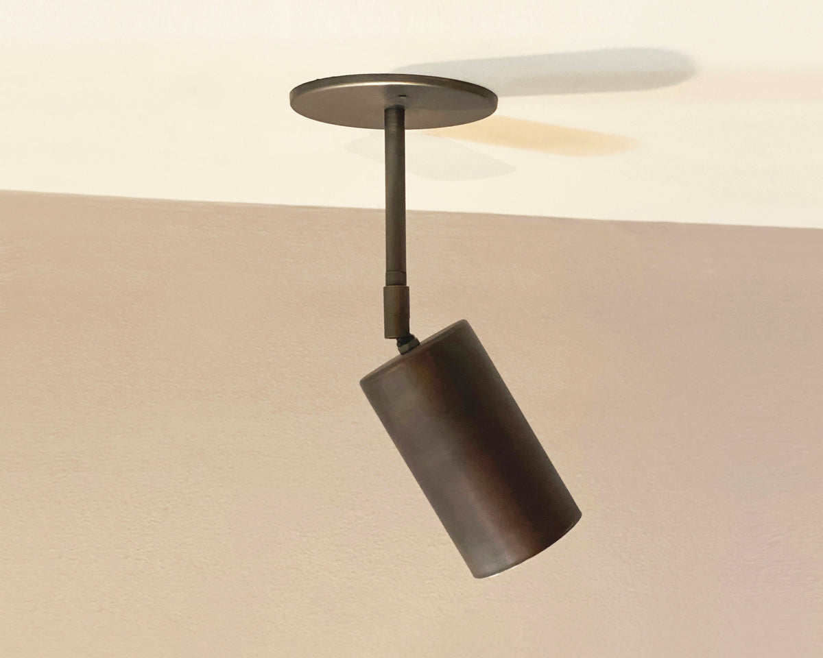 The Cove Drop Spot by Long Made Co. is a modern cylindrical solid steel downlight on a round base, angled downward against beige and off-white walls and ceiling for a sleek, minimalist spotlight effect.