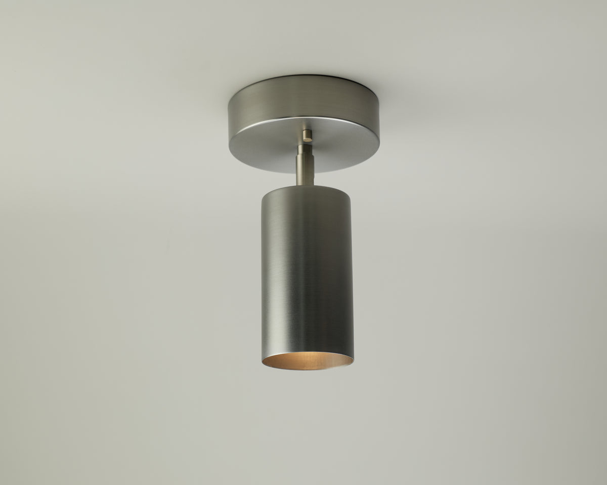 A modern Cove Adjustable Chunky Spot ceiling light fixture by Long Made Co., featuring a brushed metal monopoint design with a cylindrical shade, is mounted on a white ceiling and casts a soft downward glow.