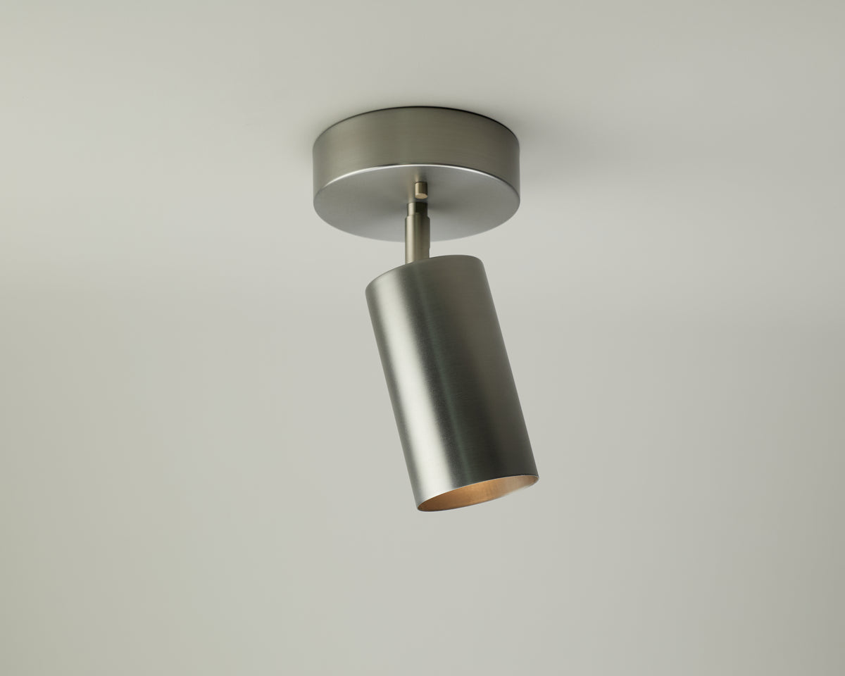 A Cove Adjustable Chunky Spot by Long Made Co., featuring a modern cylindrical brushed metal design from the Cove Series Spotlights collection, is mounted on a white ceiling with its light angled downward.