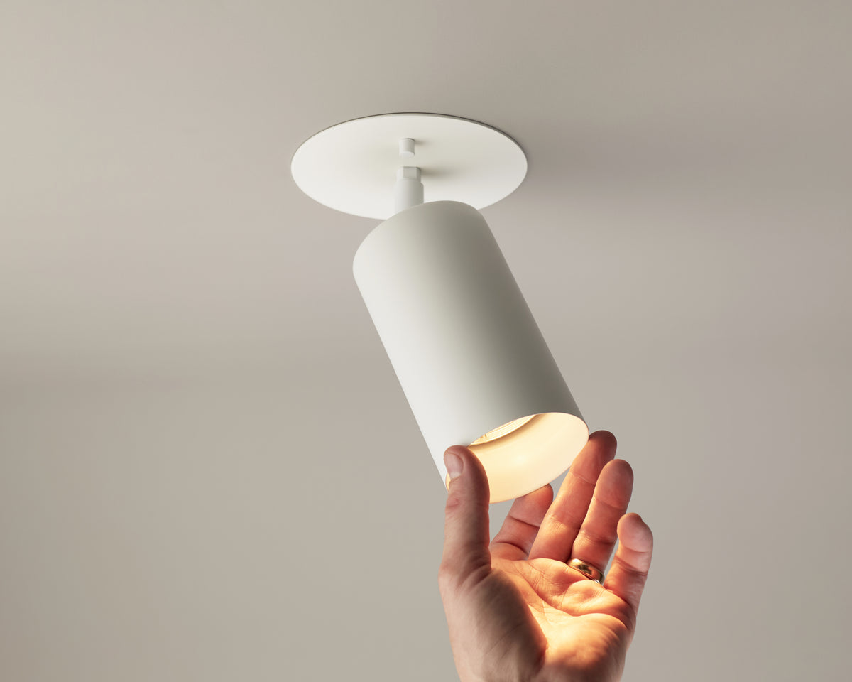 A hand adjusts the Cove Adjustable Spot by Long Made Co.—a cylindrical white ceiling downlight with a round base—casting a gentle glow against a plain, light-colored ceiling, ideal for those seeking minimalist lighting solutions.