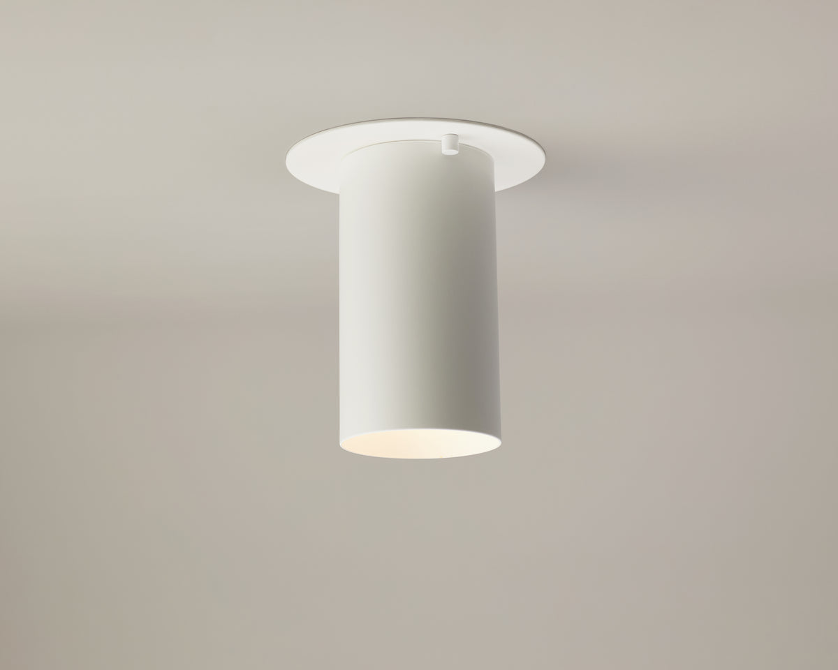 The Cove Flush Spot by Long Made Co. is a modern cylindrical white ceiling light fixture mounted on a flat, round base against a plain, light-colored ceiling and wall backdrop, offering a minimalist lighting solution and serving as an elegant alternative to traditional recessed can downlights.