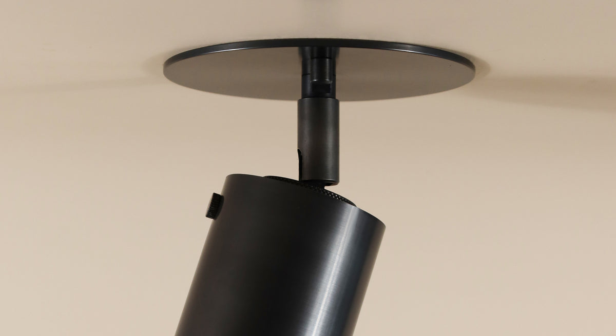 A close-up of the Long Made Co. Brass Cylinder Adjustable Spot - Top Mount, a black directional downlight fixture mounted on a white ceiling with the light angled downward.