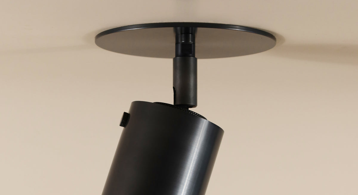 A close-up of the Long Made Co. Brass Cylinder Adjustable Spot - Top Mount, a black cylindrical directional ceiling downlight with a round base plate, is angled downward on a light-colored ceiling.