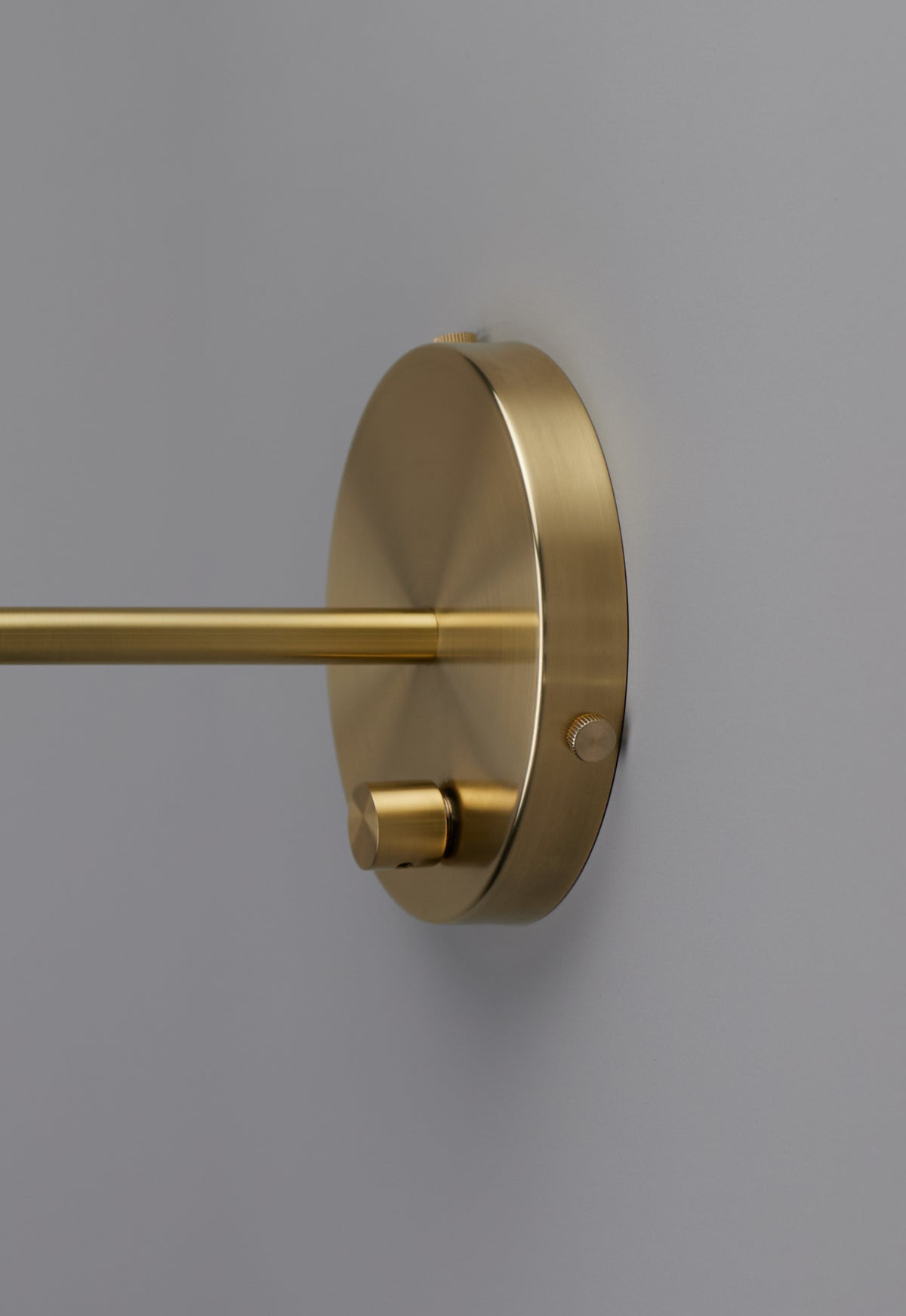 A close-up of the Highland Bent Wall Lamp by Long Made Co. shows its modern gold-colored metal wall mount and cylindrical rod set against a plain light gray wall—the sleek design echoes the signature Highland Series aesthetic, making it an ideal accent for a brass bedside lamp.
