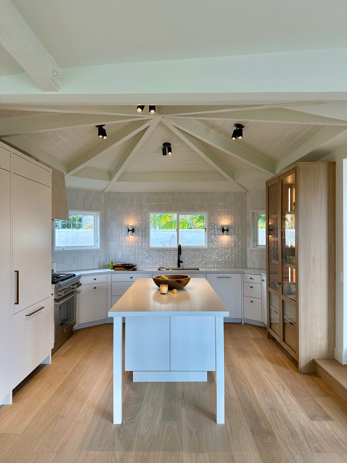 Modern kitchen with light wood floors, white cabinets, a central island, stainless steel appliances, and large windows. The ceiling features exposed beams and three Long Made Co. Brass Cylinder Triple Spots for optimal illumination.