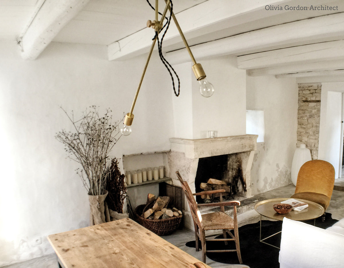 A cozy, rustic living room with white walls, exposed beams, a stone fireplace, wooden chairs, a round coffee table, and the Task Articulating Chandelier by Long Made Co. overhead. Dried branches in a vase sit by the fireplace.
