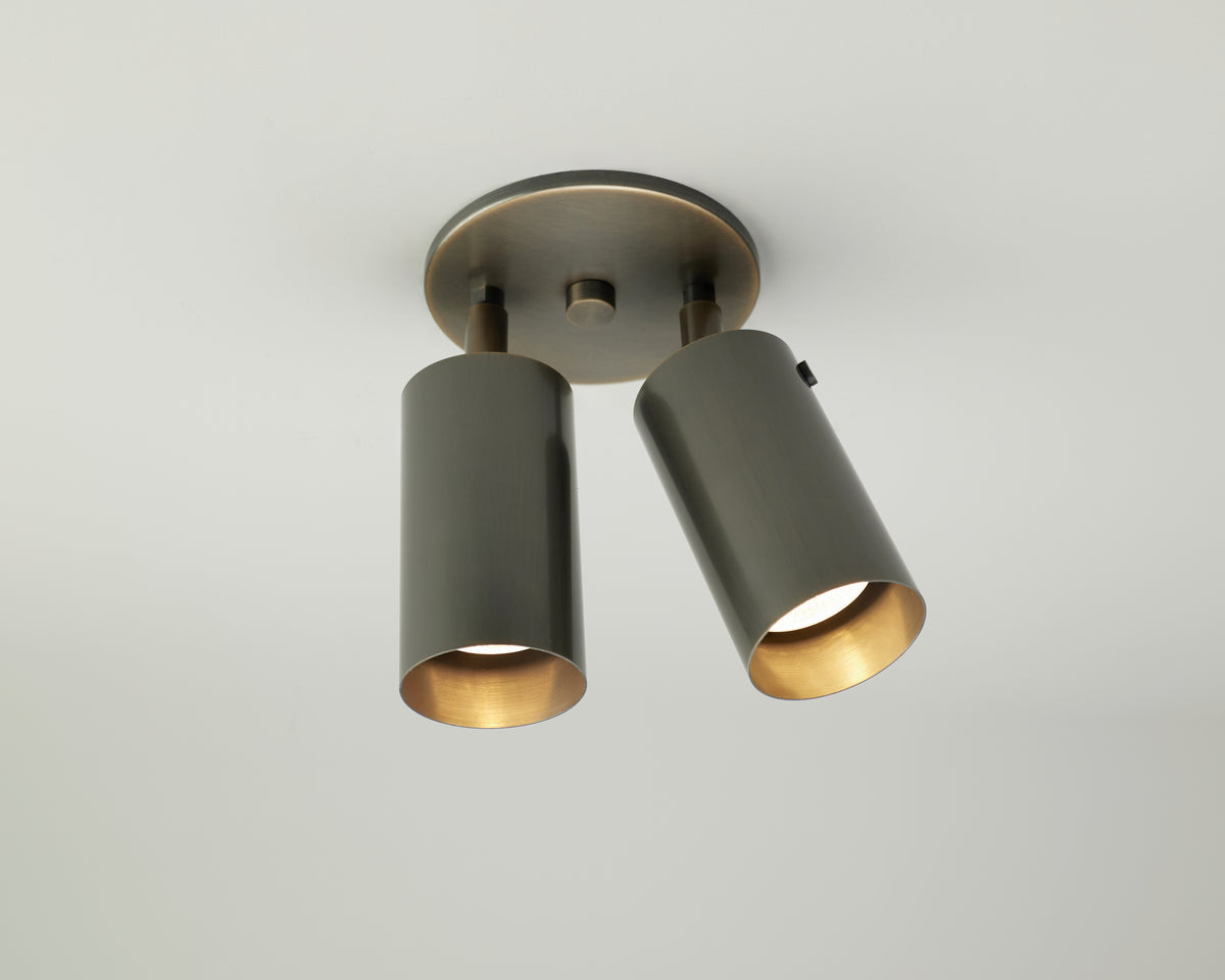 The Brass Cylinder Double Spot by Long Made Co. is a modern ceiling light featuring two cylindrical metallic fixtures in a brass finish, each attached to a round base and mounted on a white ceiling. This adjustable spotlight allows both fixtures to be directed downward and angled apart for versatile illumination, showcasing a sleek mid-century modern style.