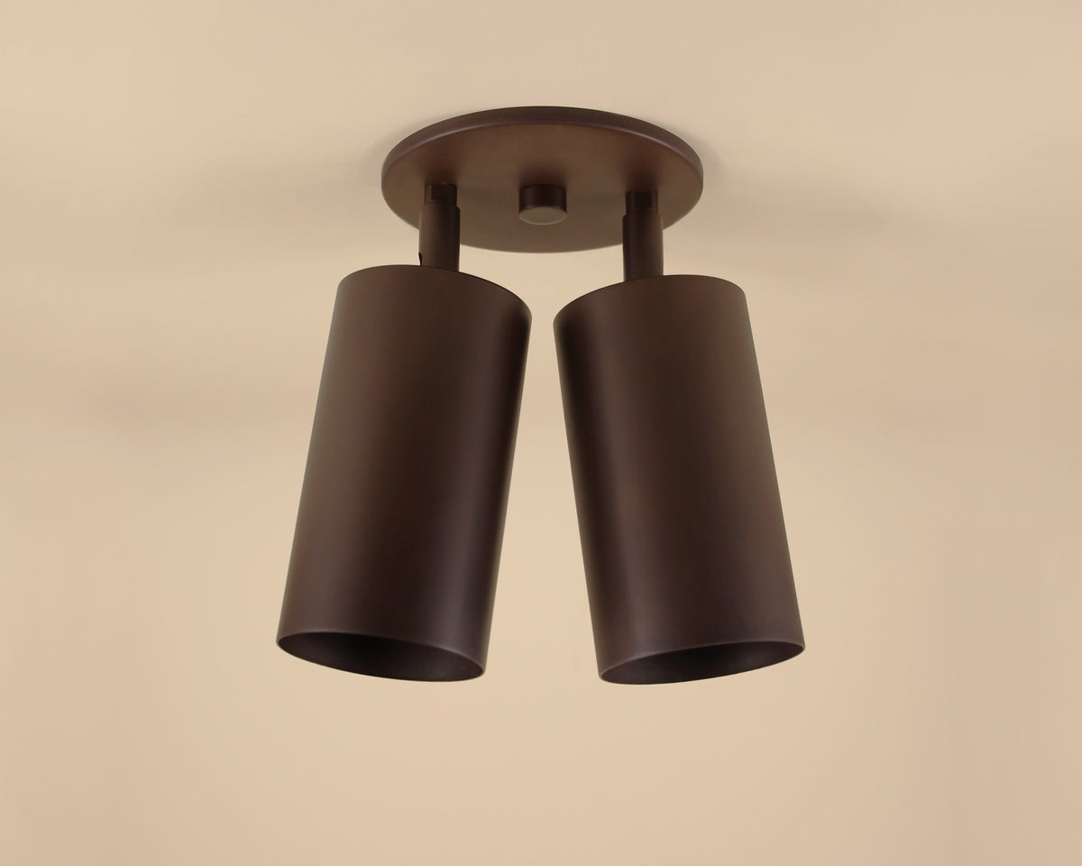 The Cove Double Spot by Long Made Co. is a modern ceiling downlight with two cylindrical matte dark brown shades on a round base, ideal for minimalist lighting on beige ceilings in any space.