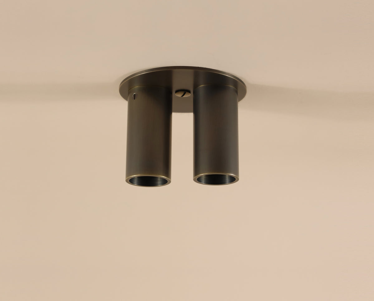 The Long Made Co. Micro Flush Spot - Double is a modern flush mount fixture featuring two cylindrical dark metal tubes side by side on a round base, ideal for ceiling installation.