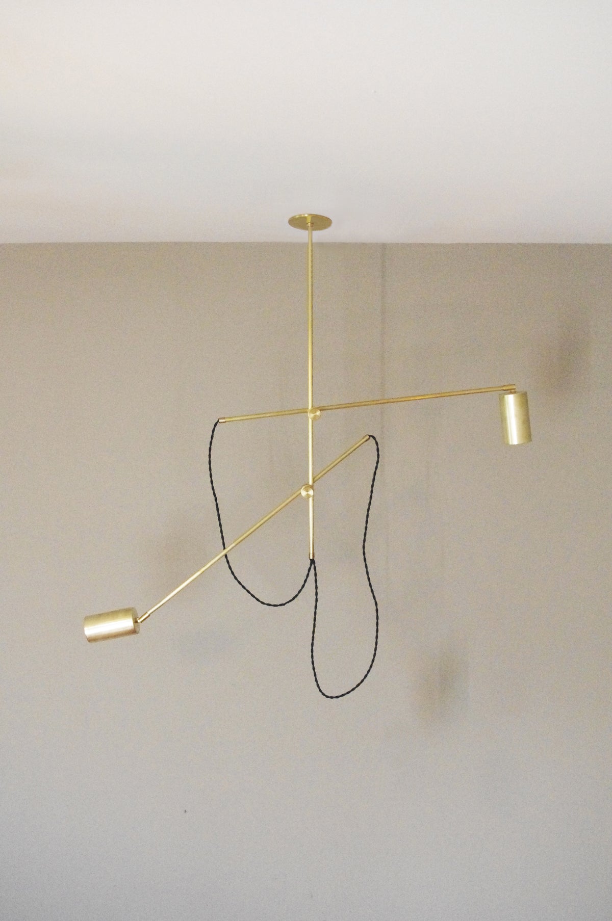 The Task Articulating Chandelier by Long Made Co. features two solid brass cylindrical lamps and a black cord, hanging from a white ceiling against a beige wall—an ideal blend of style and function for versatile task lighting.