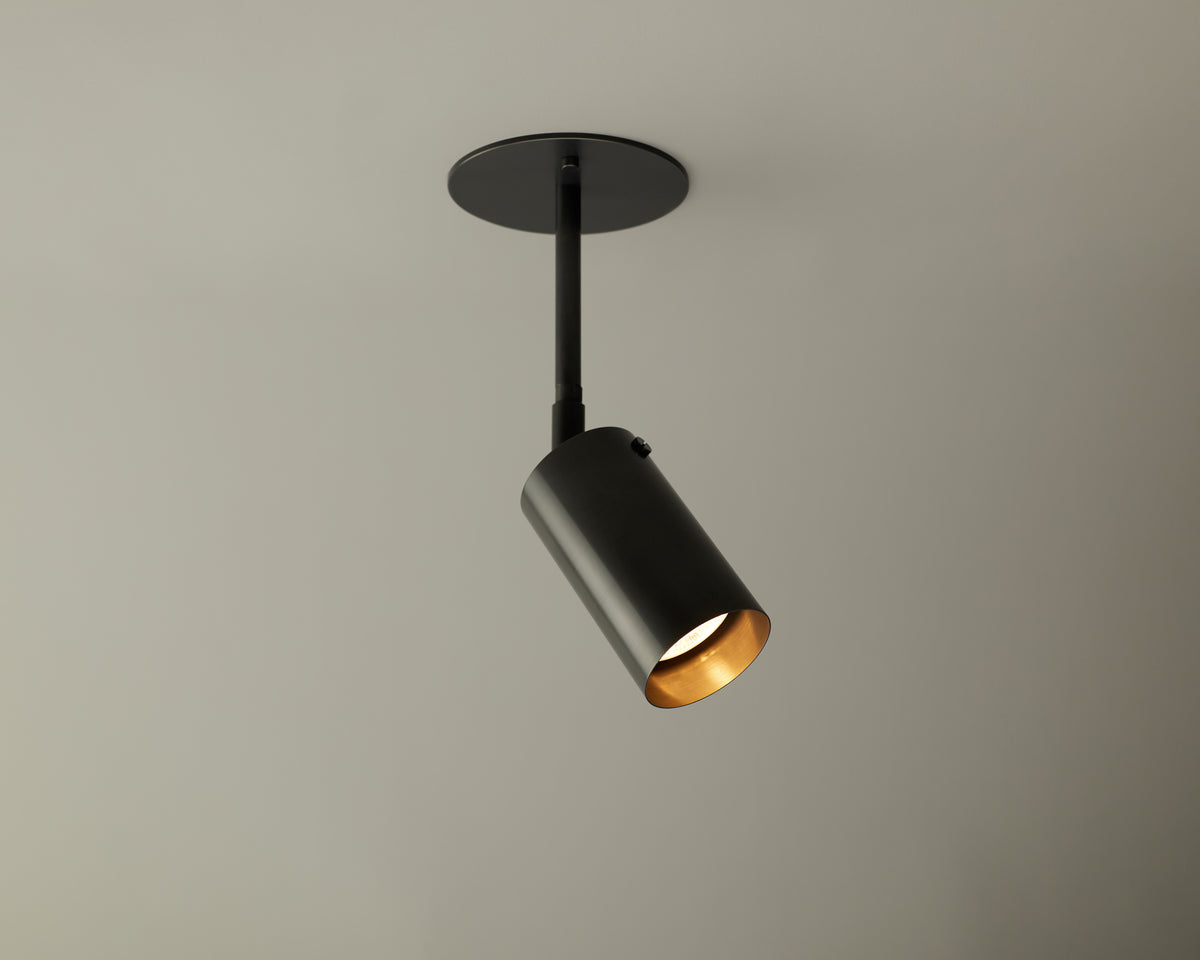 The Brass Cylinder Drop Spot - Top Mount by Long Made Co. is a modern black ceiling light mounted on a light gray ceiling, angling its beam downward and to the side for focused illumination with sleek mid-century modern style.