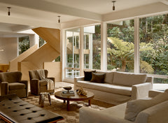A modern living room with large windows, neutral sofas, a round wooden coffee table, and a light wood staircase features Long Made Co.'s Brass Cylinder Drop Spot - Top Mount for a mid-century modern lighting accent in the cozy, sunlit space.