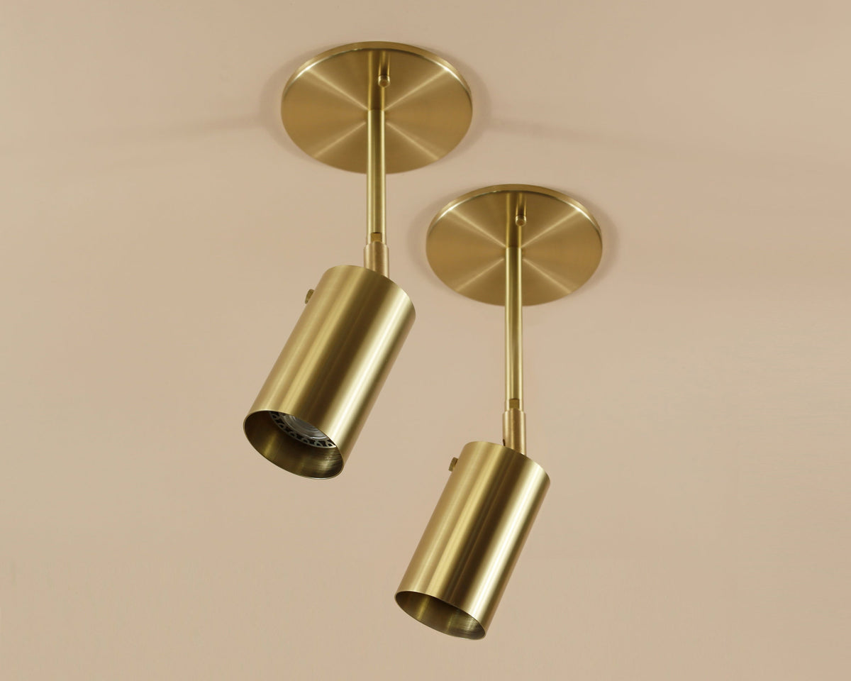 Two Brass Cylinder Drop Spot - Top Mount lights by Long Made Co. feature modern cylindrical shades in brass, mounted on a beige ceiling and angled downward in different directions for stylish, directional illumination.
