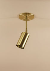 The Brass Cylinder Drop Spot - Top Mount by Long Made Co. is a brass-colored, cylindrical ceiling spotlight pictured angled downward from a round ceiling plate against a plain beige background.