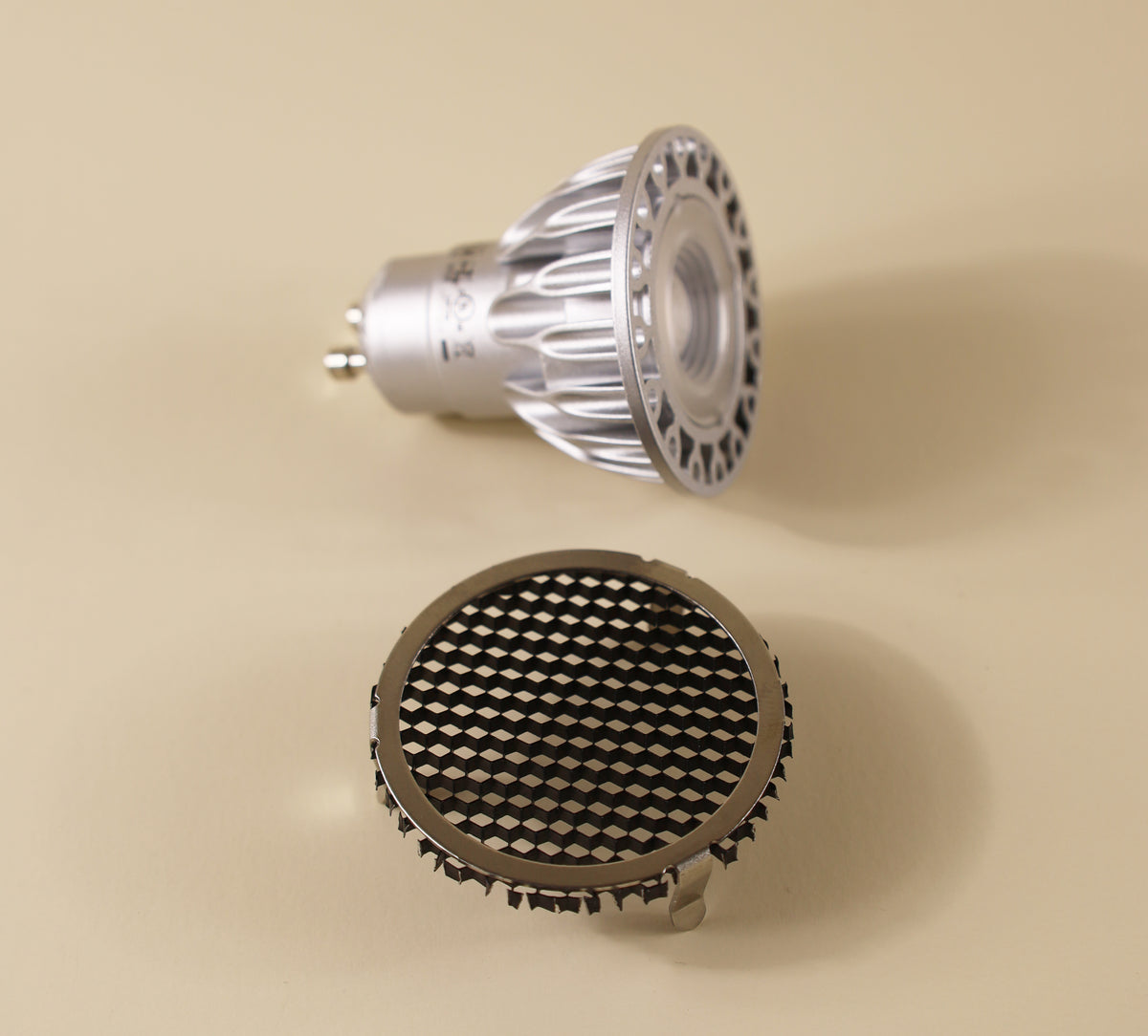 A silver MR16 GU10 LED spotlight bulb and a detached Long Made Co. Louver - EmeryAllen - MR16 accessory featuring a crisscross honeycomb pattern for glare reduction are placed on a beige surface.