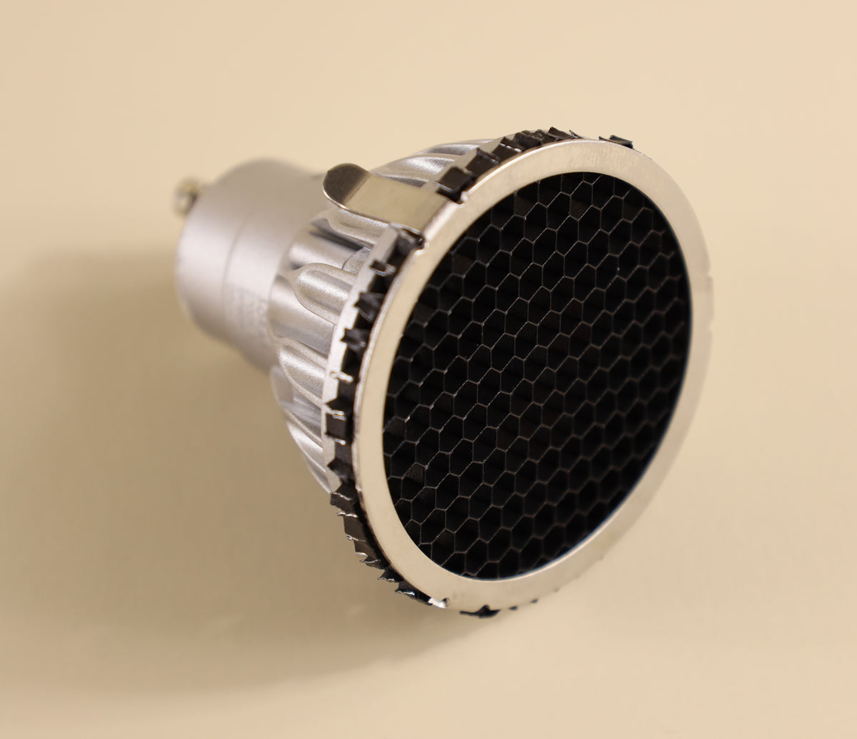 A close-up of the Long Made Co. Louver – EmeryAllen – MR16, a silver LED MR16 GU10 spotlight bulb featuring a black honeycomb louver grid filter on the lens for effective glare reduction, placed on a beige surface.