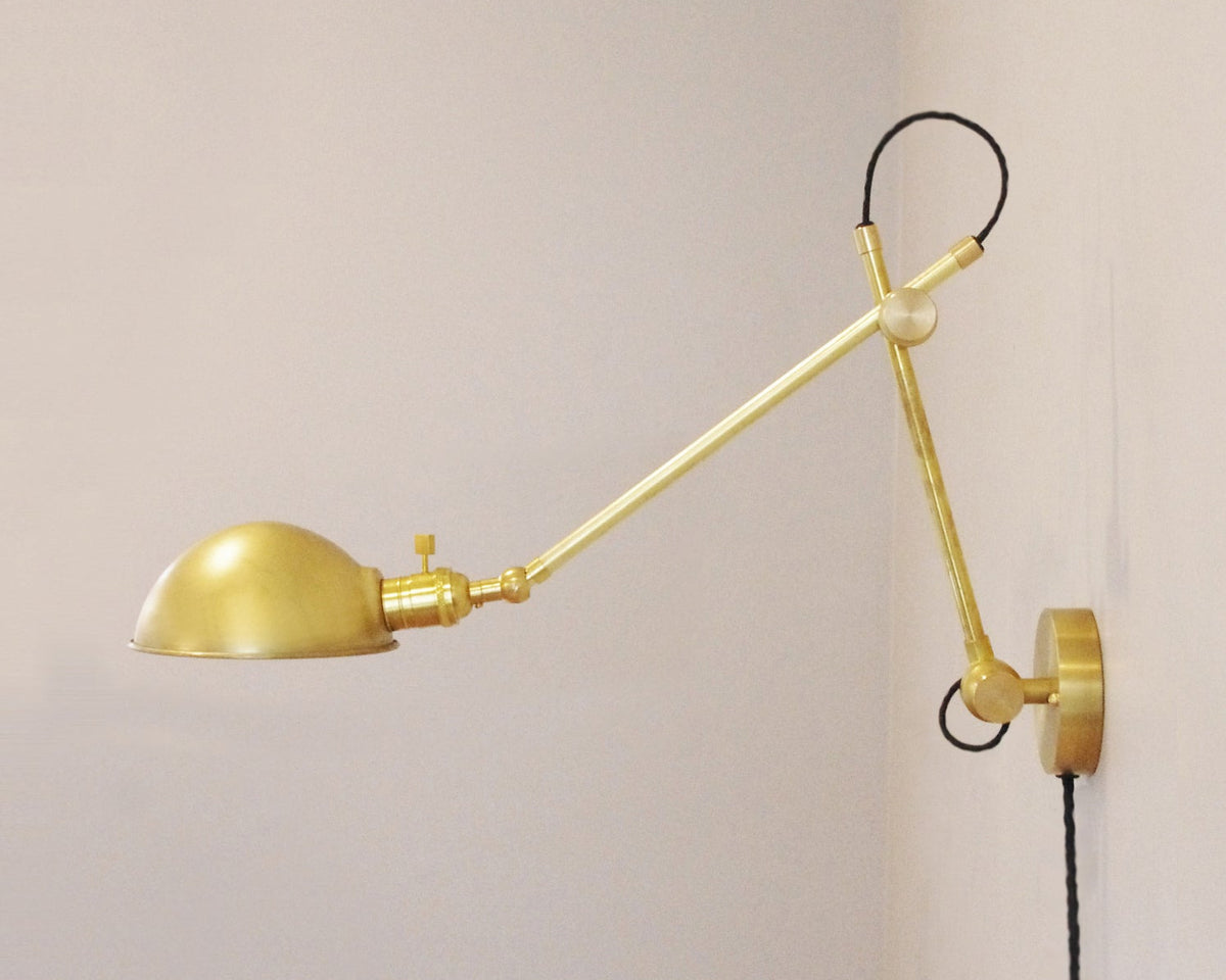 The Task Articulating Wall Lamp by Long Made Co. is a modern brass wall sconce with a dome-shaped shade, adjustable joints, and a black cord extending from the mounting plate, shown attached to a white wall.