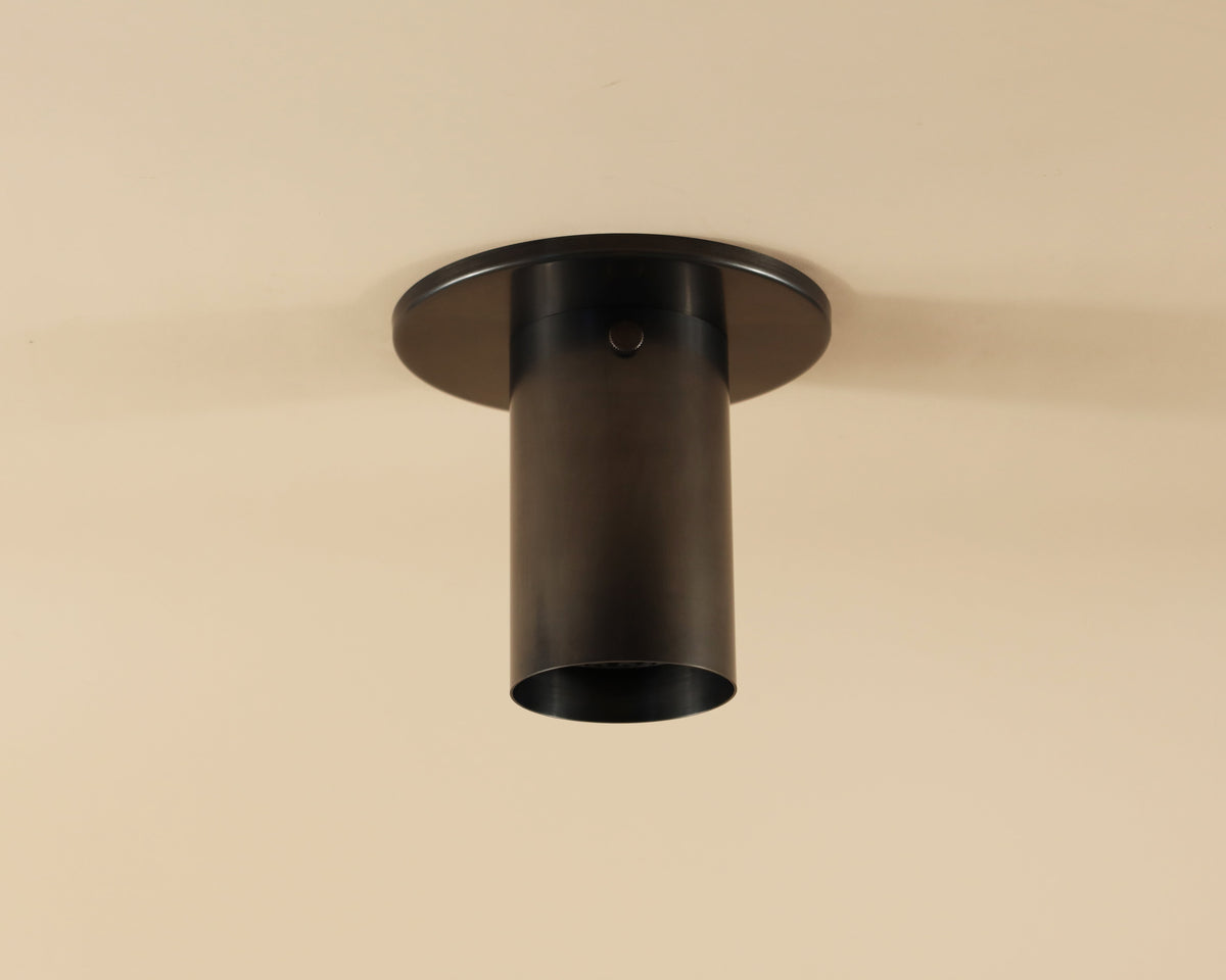 The Brass Cylinder Flush Spot by Long Made Co. is a modern, cylindrical black ceiling light with a minimalist design, inspired by mid-century modern style, and is elegantly mounted on a beige ceiling.