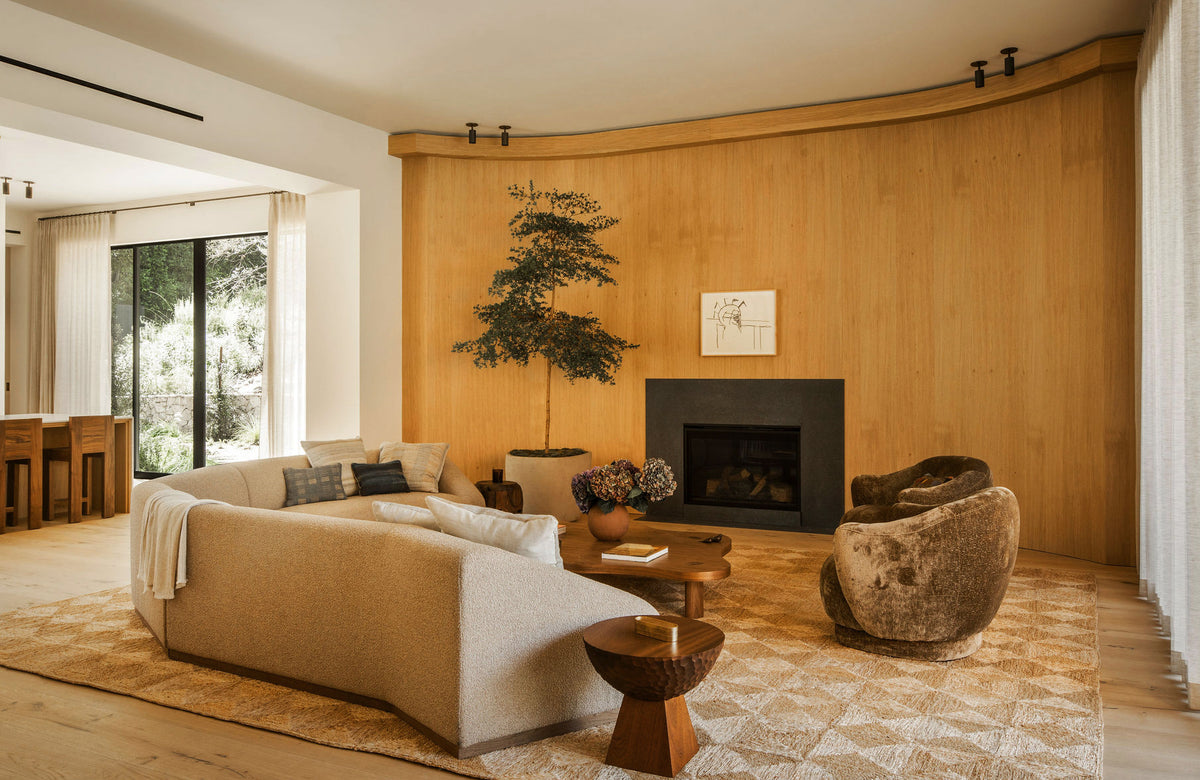 A cozy living room with light wood walls, fireplace, modern furniture, a Long Made Co. Brass Cylinder Adjustable Spot - Top Mount ceiling light, large rug, beige sofa, brown armchair, coffee table, indoor plant, and sheer curtains filtering sunlight.