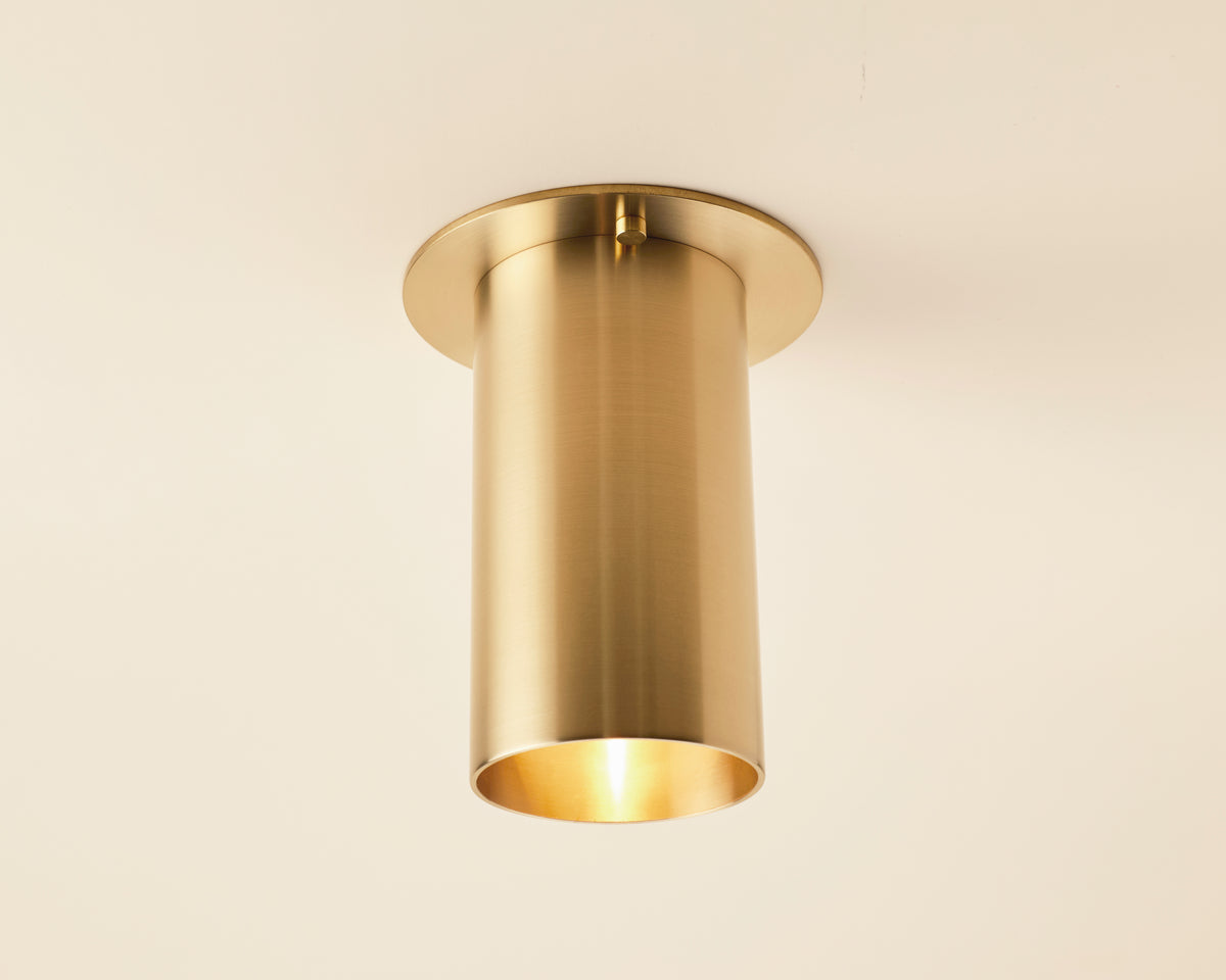 The Highland Flush Spot by Long Made Co. is a modern brass cylinder light with a warm downward glow, mounted on a smooth, light ceiling—ideal for those desiring elegant flush monopoint lighting.