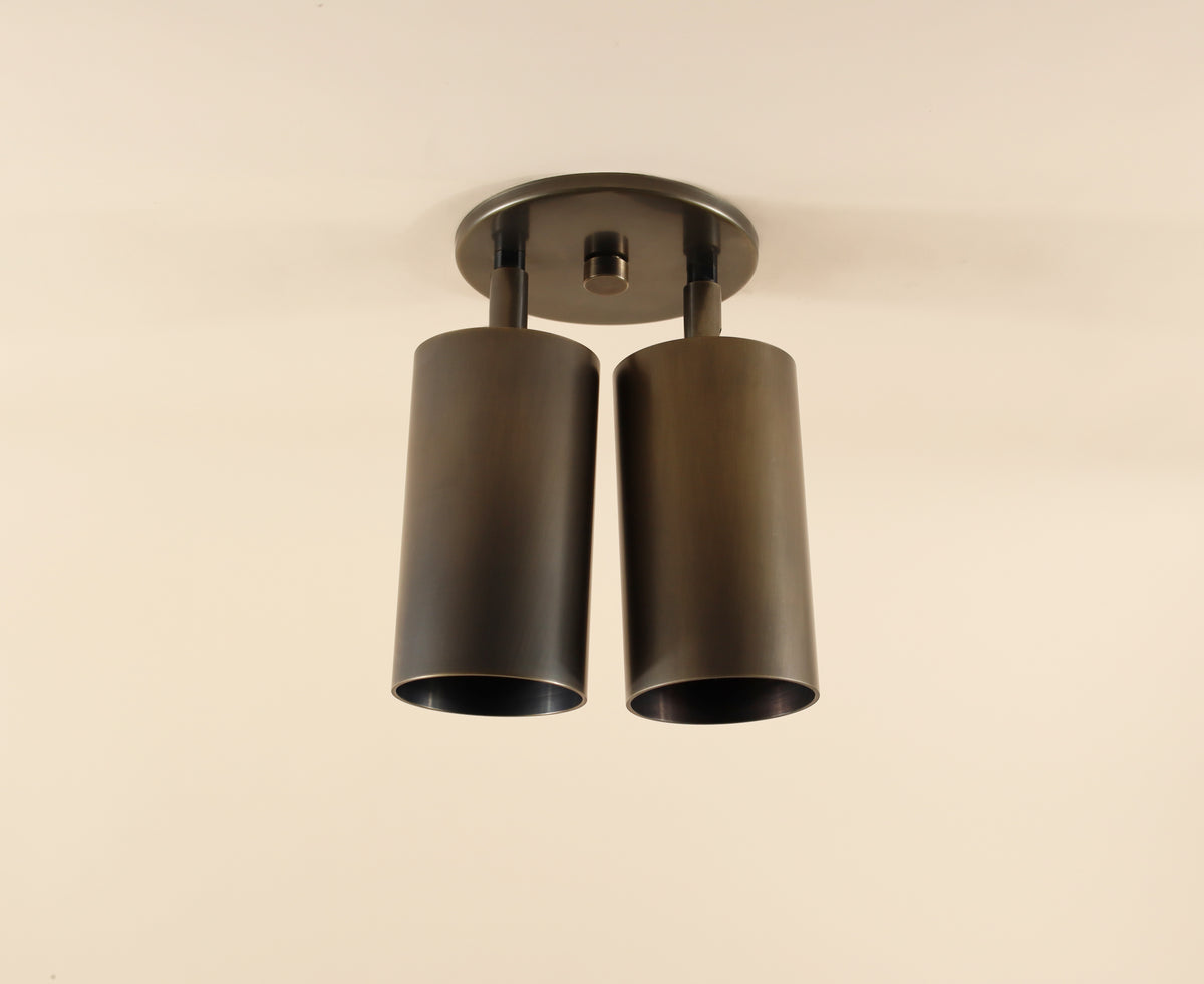 The Highland Double Spot by Long Made Co. is a modern ceiling fixture with two cylinder lights in a dark finish on a round base—ideal minimalist lighting for any space.