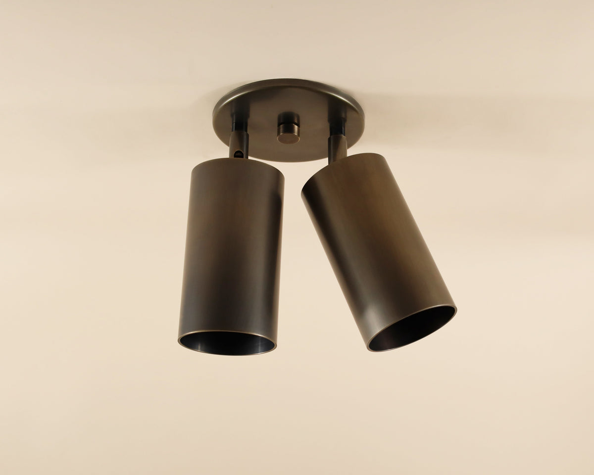 The Highland Double Spot by Long Made Co. features two cylindrical black shades, slightly angled downward on a round base—ideal for minimalist lighting against a light ceiling.