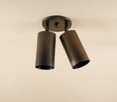 A close-up of the Highland Double Spot by Long Made Co.—a modern ceiling light with two adjustable black cylinder lights on a round base—showcases its minimalist design against a beige ceiling.