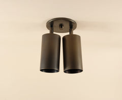 The Highland Double Spot by Long Made Co. is a modern ceiling fixture with two brushed metal cylinder lights on a round base, ideal for minimalist interiors.