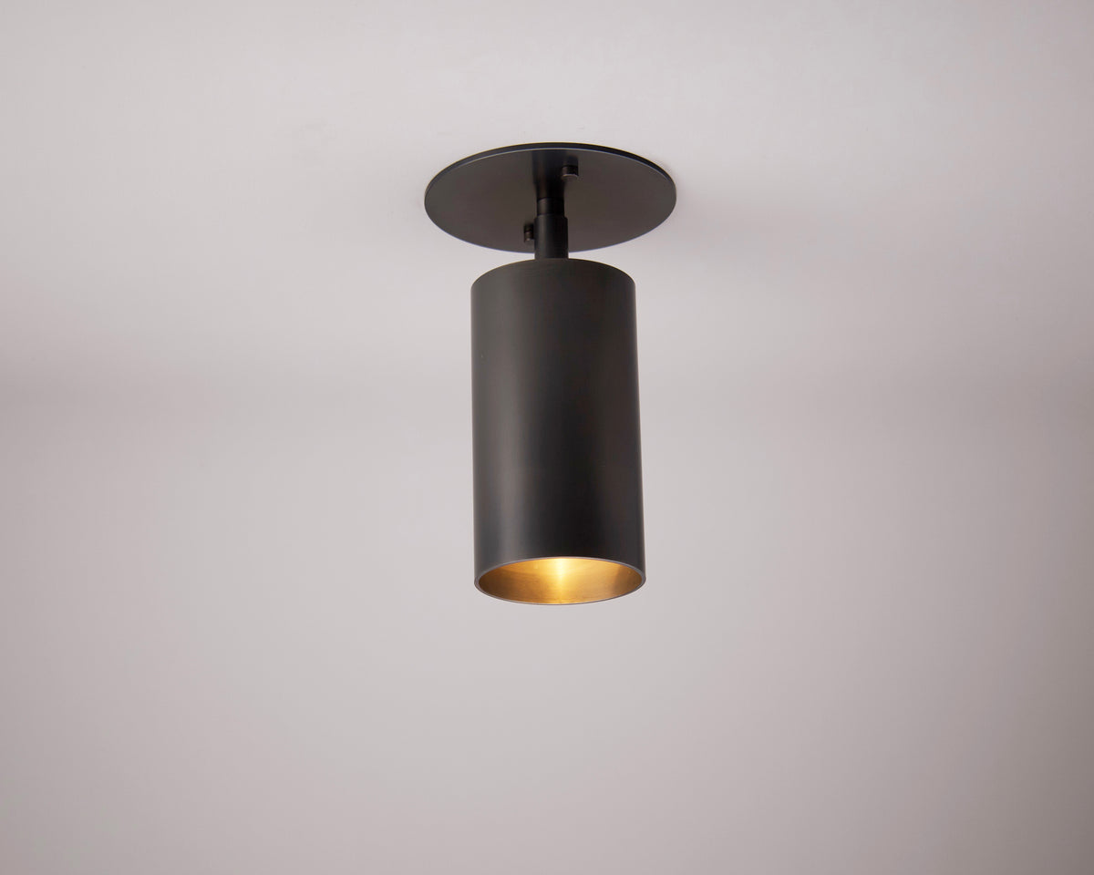The Highland Adjustable Spot by Long Made Co. is a modern, minimalist black cylindrical fixture that casts a warm glow, elegantly mounted on a white ceiling.