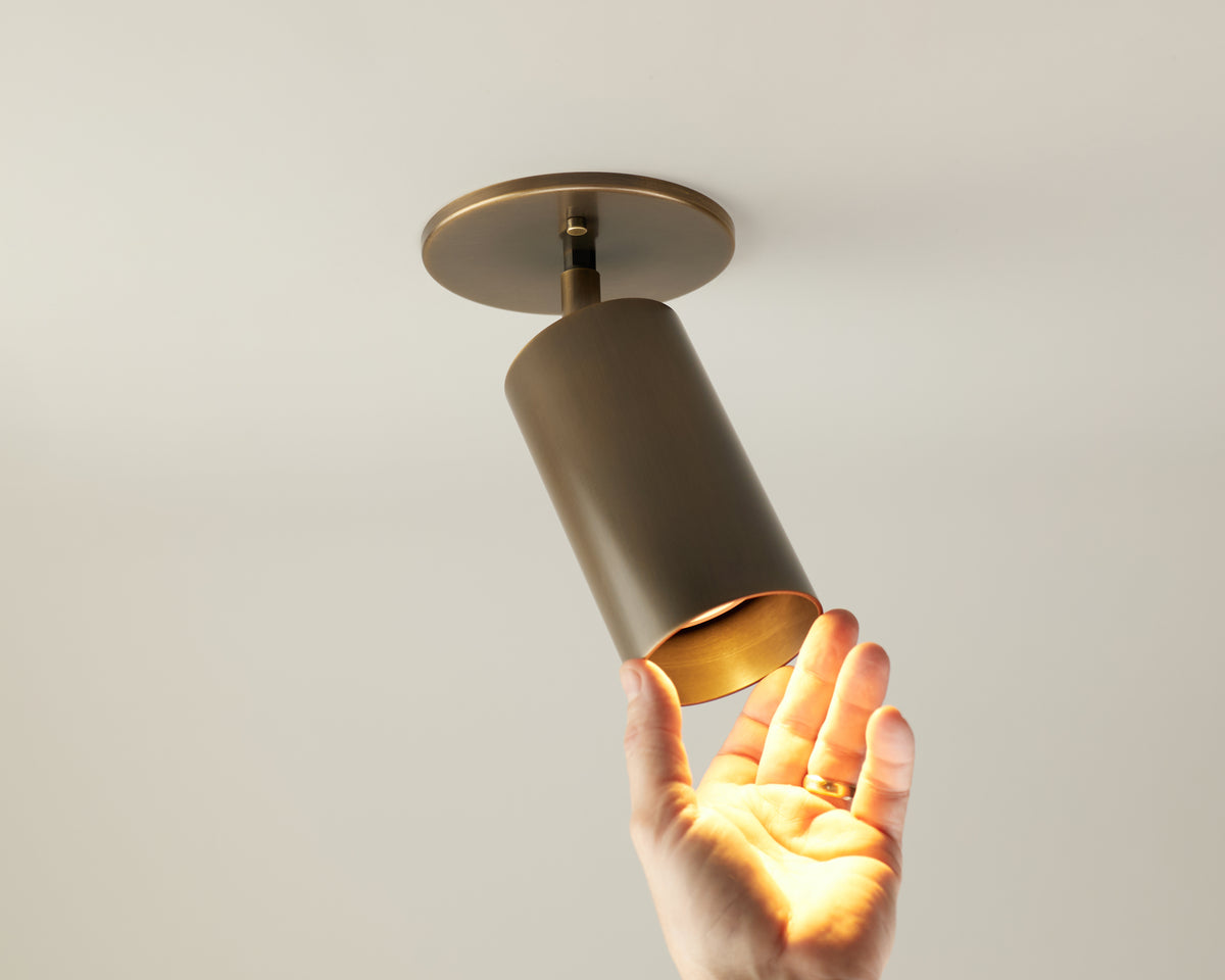 A hand reaches up toward the ceiling-mounted Highland Adjustable Spot by Long Made Co., its minimalist cylinder design casting a warm glow against a plain white ceiling—perfect for sophisticated residential and commercial lighting schemes.