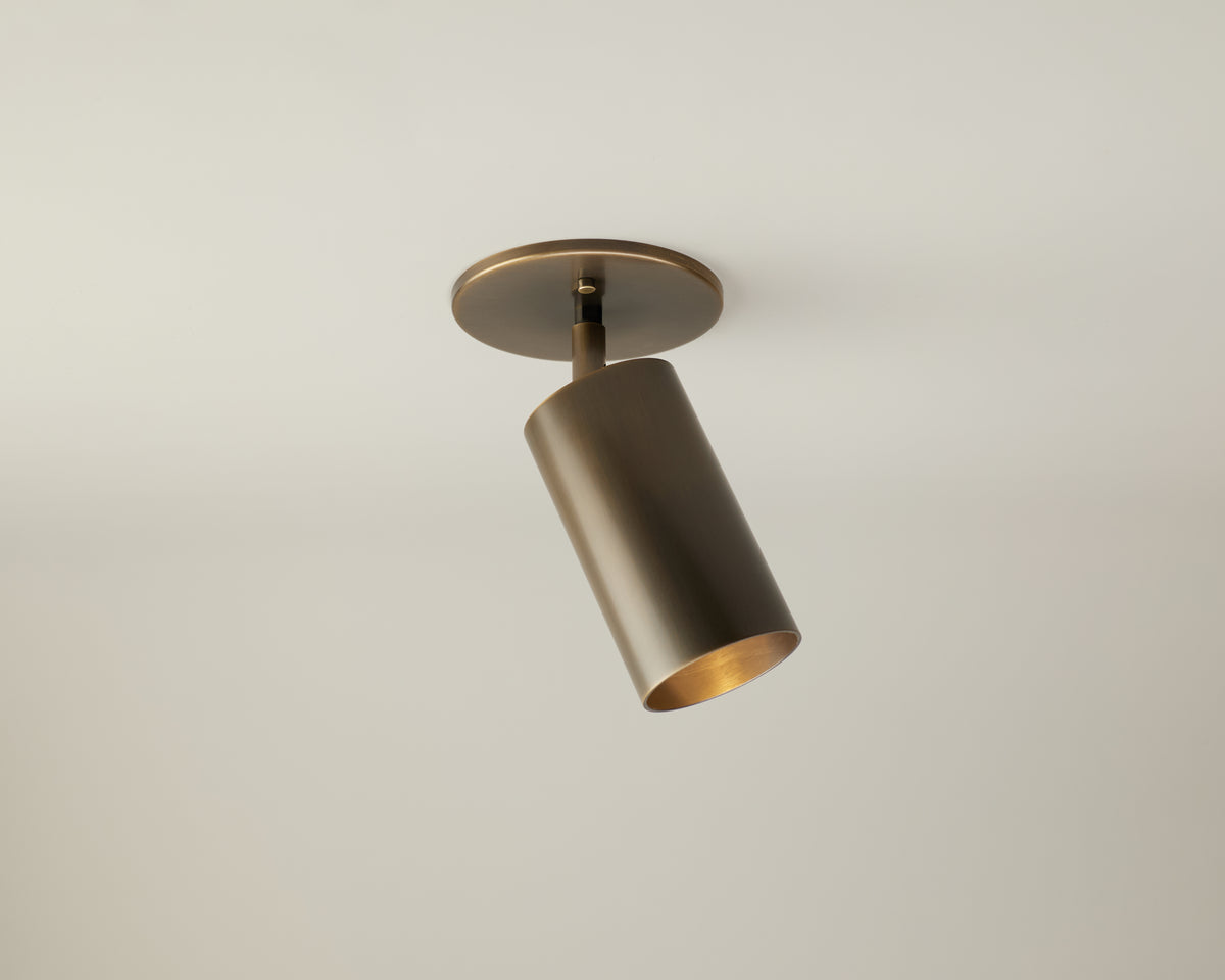 The Highland Adjustable Spot by Long Made Co. is a modern, cylindrical brass ceiling spotlight mounted on a white ceiling and angled downward for directional lighting. Perfect for both residential and commercial spaces, this minimalist fixture exemplifies the elegance of handmade brass spotlights while delivering focused illumination.