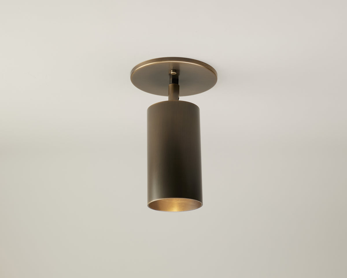The Highland Adjustable Spot by Long Made Co. is a modern, minimalist brass cylinder light fixture mounted on a white ceiling, emitting focused downward light—perfect for both residential and commercial lighting settings.