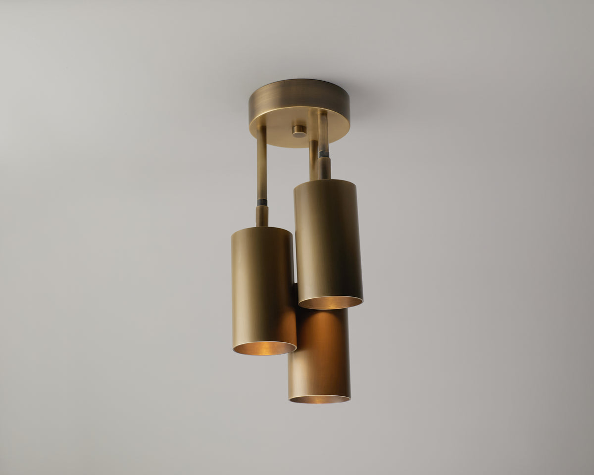 The Highland Cascade Drop Spot by Long Made Co. is a modern brass ceiling light fixture featuring three handmade, heavy brass cylinder lights at varying heights, elegantly suspended against a plain white ceiling. Combining contemporary style with artisanal craftsmanship, this piece creates a striking focal point in any space.