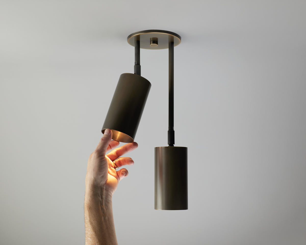 A modern two-light cylinder pendant ceiling fixture in a dark bronze finish, shown with a hand adjusting one of the shades against a plain light gray ceiling.