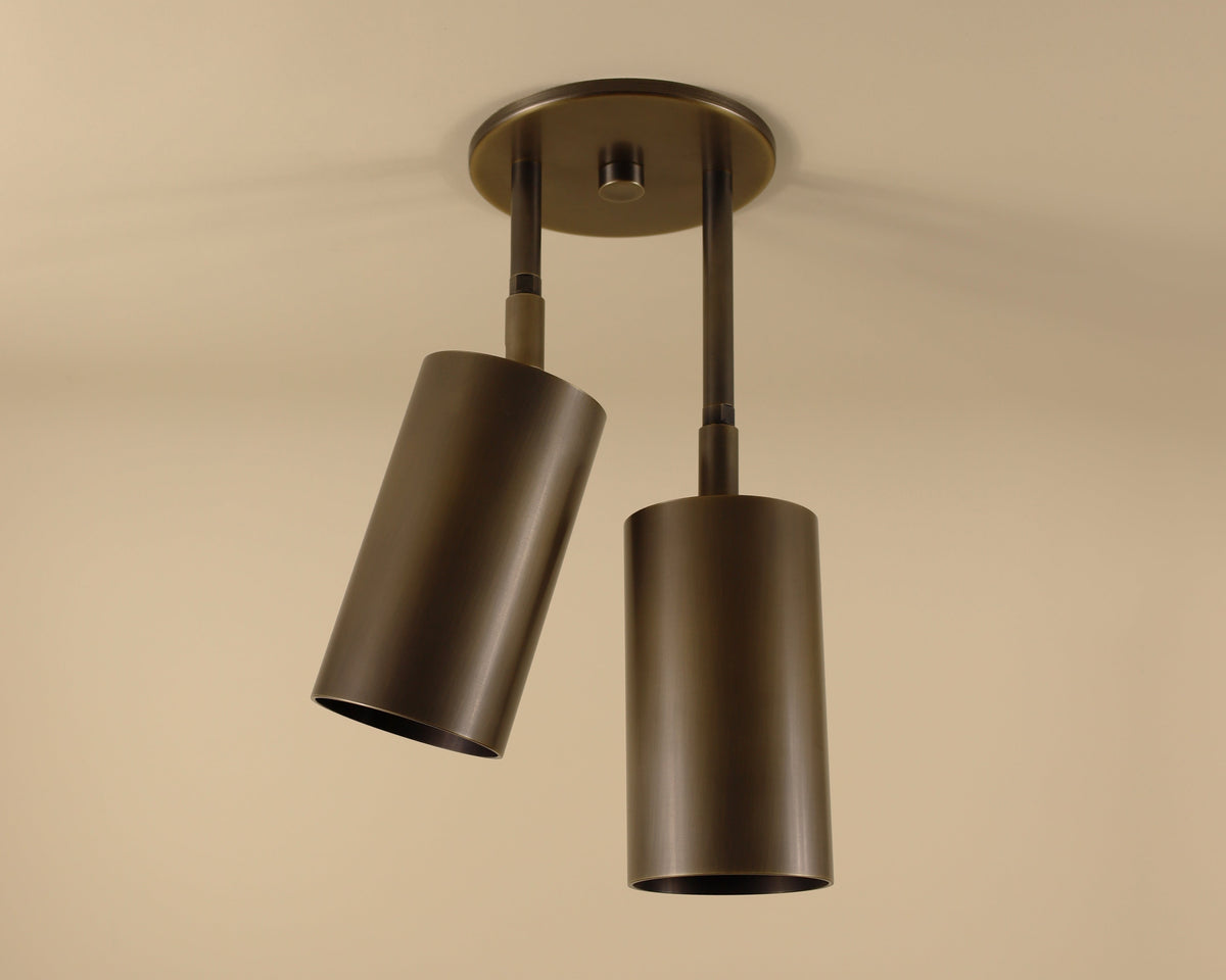 The Highland Double Drop Spot by Long Made Co. features two handmade cylinder lights on a round base, each angled uniquely to cast a warm glow against the neutral beige ceiling.