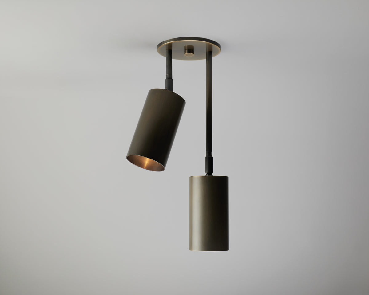 Two Highland Double Drop Spot pendant lights by Long Made Co. with a modern, minimalist cylindrical design hang from a round ceiling mount against a plain white background. These adjustable cylinder lights provide versatility—one light is tilted at an angle while the other hangs straight down.