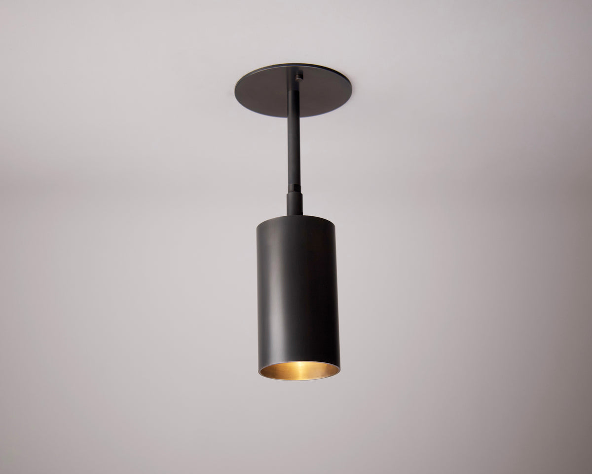 The Highland Drop Spot by Long Made Co. is a modern black cylinder light hanging from a white ceiling, casting a soft warm glow below—perfect for adding elegant, directional lighting to contemporary interiors.