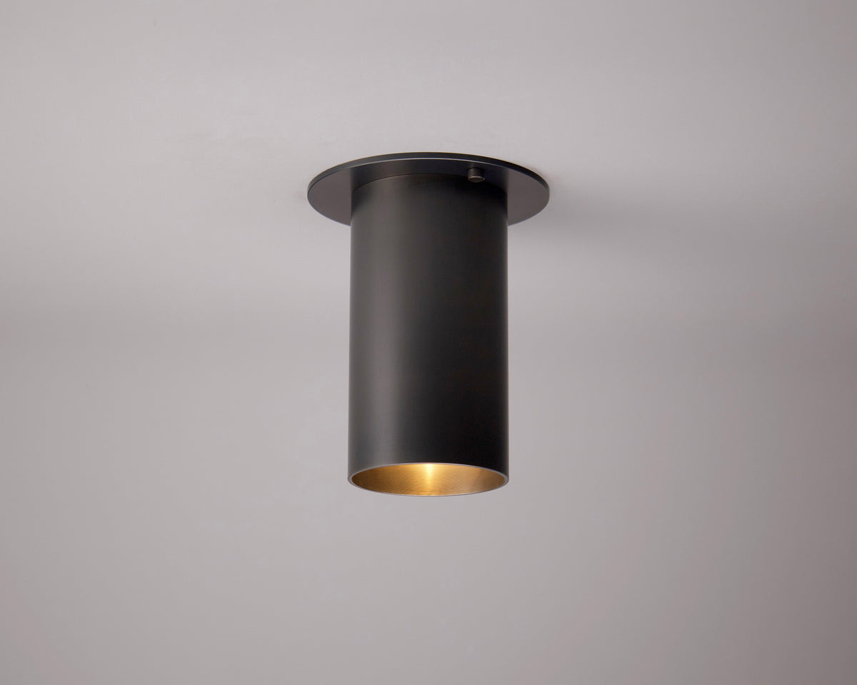The Highland Flush Spot by Long Made Co. is a black cylindrical ceiling fixture with a gold interior, flush-mounted on a white ceiling. Inspired by heavy brass lights, it hangs straight down and emits warm light from its open bottom.