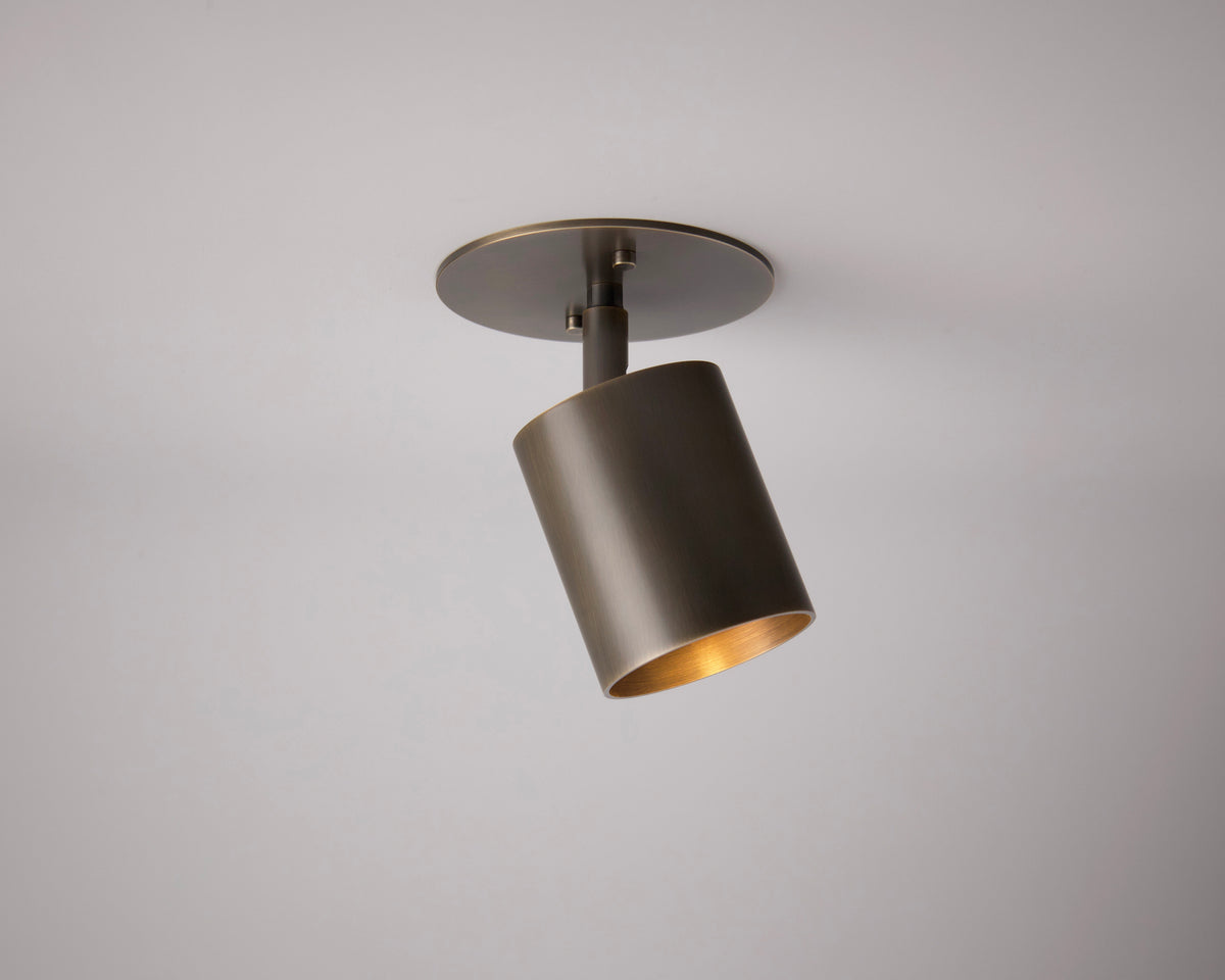 The Long Made Co. Highland Adjustable Spot - Stocky is a modern, cylindrical brass spotlight on a round base with a bronze finish, angled downward on a white ceiling for focused illumination.