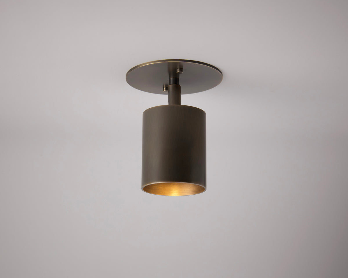 The Highland Adjustable Spot - Stocky by Long Made Co. is a modern cylindrical bronze ceiling light with a flat round base, mounted on a white ceiling and casting warm light downward—a perfect minimalist brass fixture.
