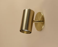 The Highland Wall Lamp w/ Switch by Long Made Co. is a modern, cylindrical brass sconce with a brushed finish, mounted on a light beige wall and angled downward for reading.