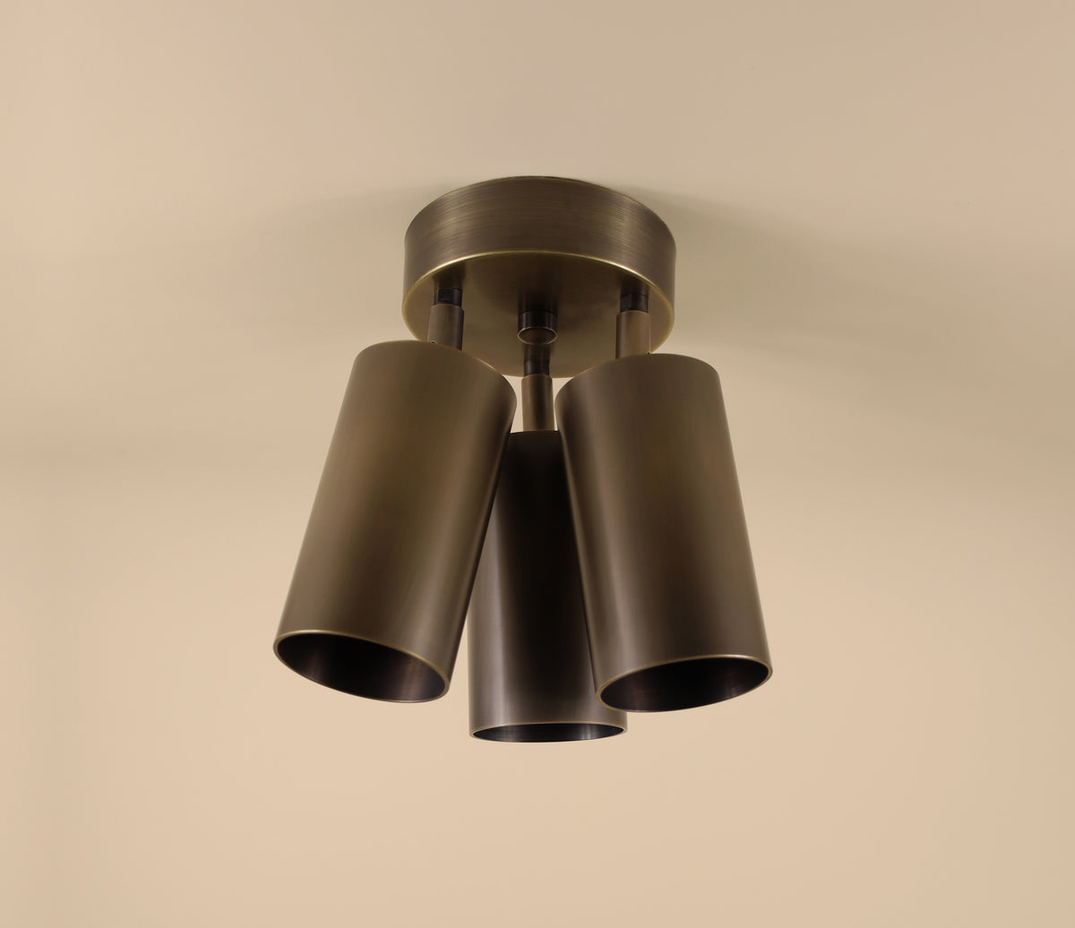 The Highland Double / Triple Spot by Long Made Co. is a modern ceiling fixture with three dark metal cylinder lights in a triangular layout, mounted on a round base against a beige ceiling.