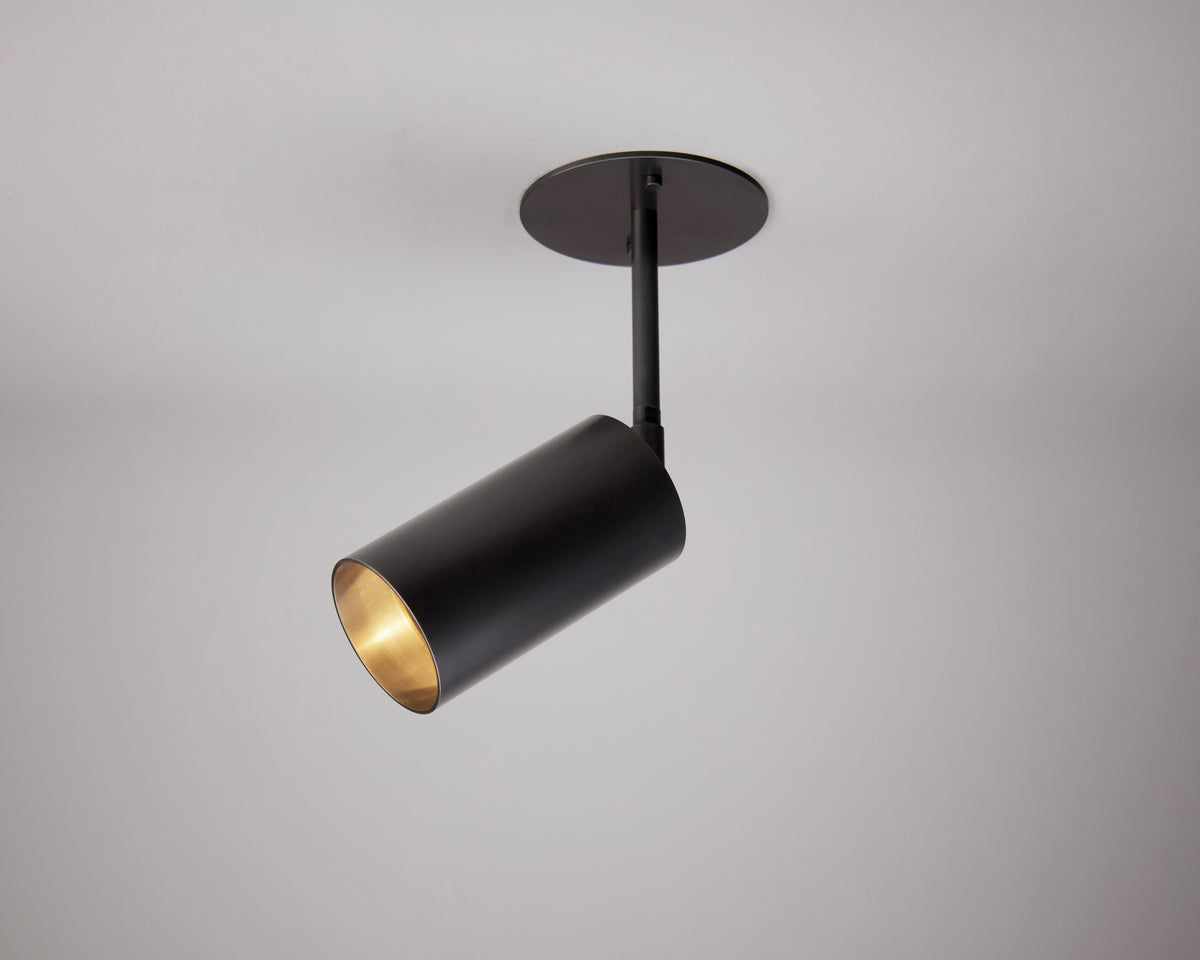 The Highland Drop Spot by Long Made Co. is a modern black cylindrical ceiling mono point spotlight with a matte finish and round base, mounted on a white ceiling. Its downward, angled light is ideal for focused task lighting.