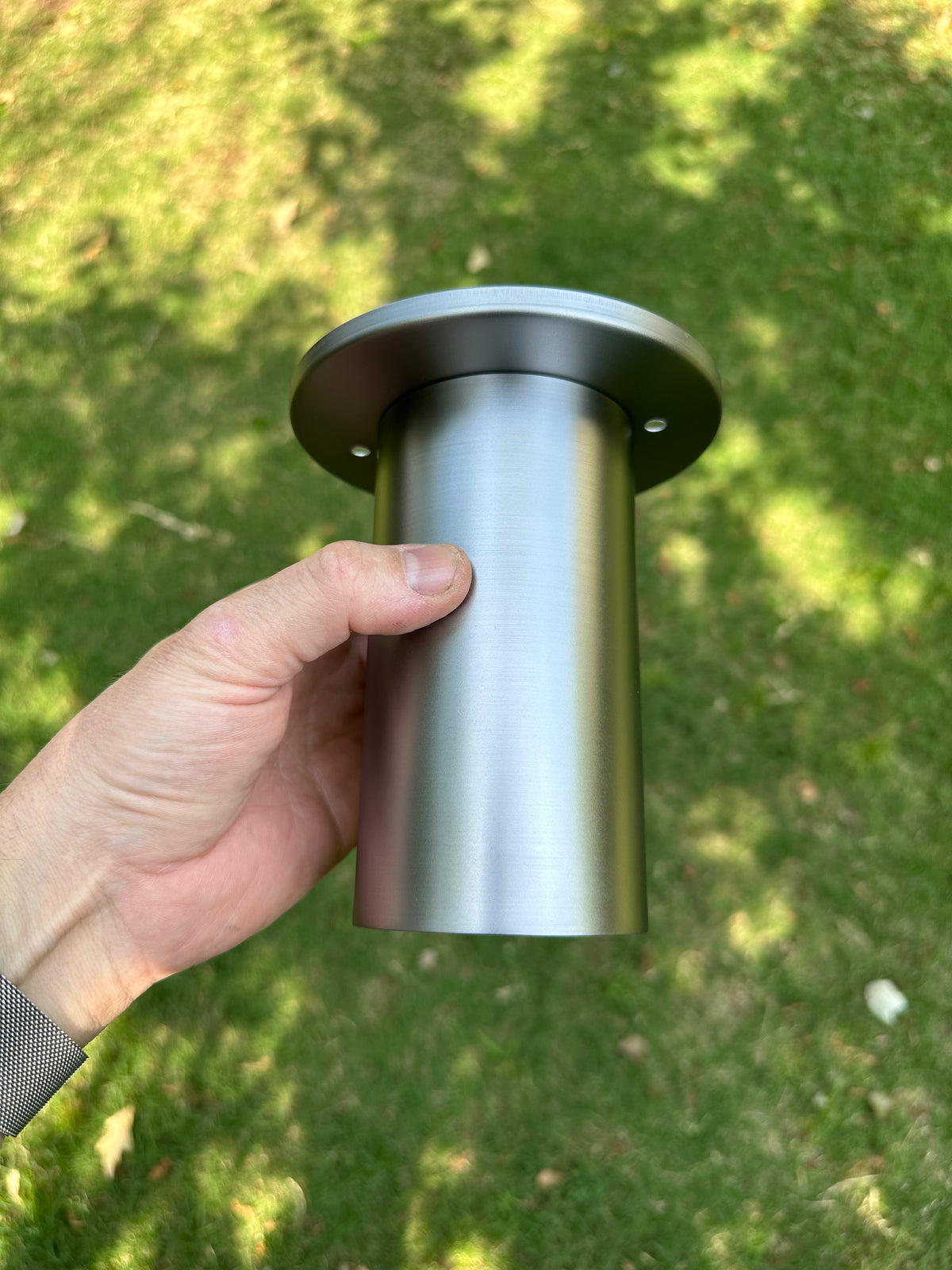 A hand holds a Long Made Co. Cove Flush Spot downlight with a circular flange outdoors, above sun-dappled grass.